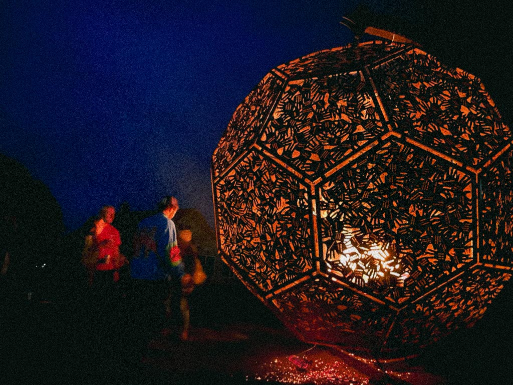 At night, a glowing large steel art sculpture with pentagon and laser-cut hands details, the InnerSun. A group of participants stand nearby, lit by the glow.