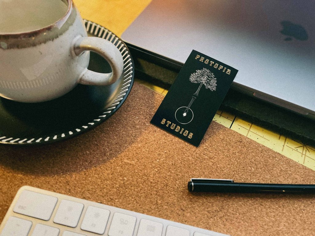 A close-up of a business card that says Protopia Studios, next to a laptop, keyboard and coffee mug. We offer writing services and creative development.