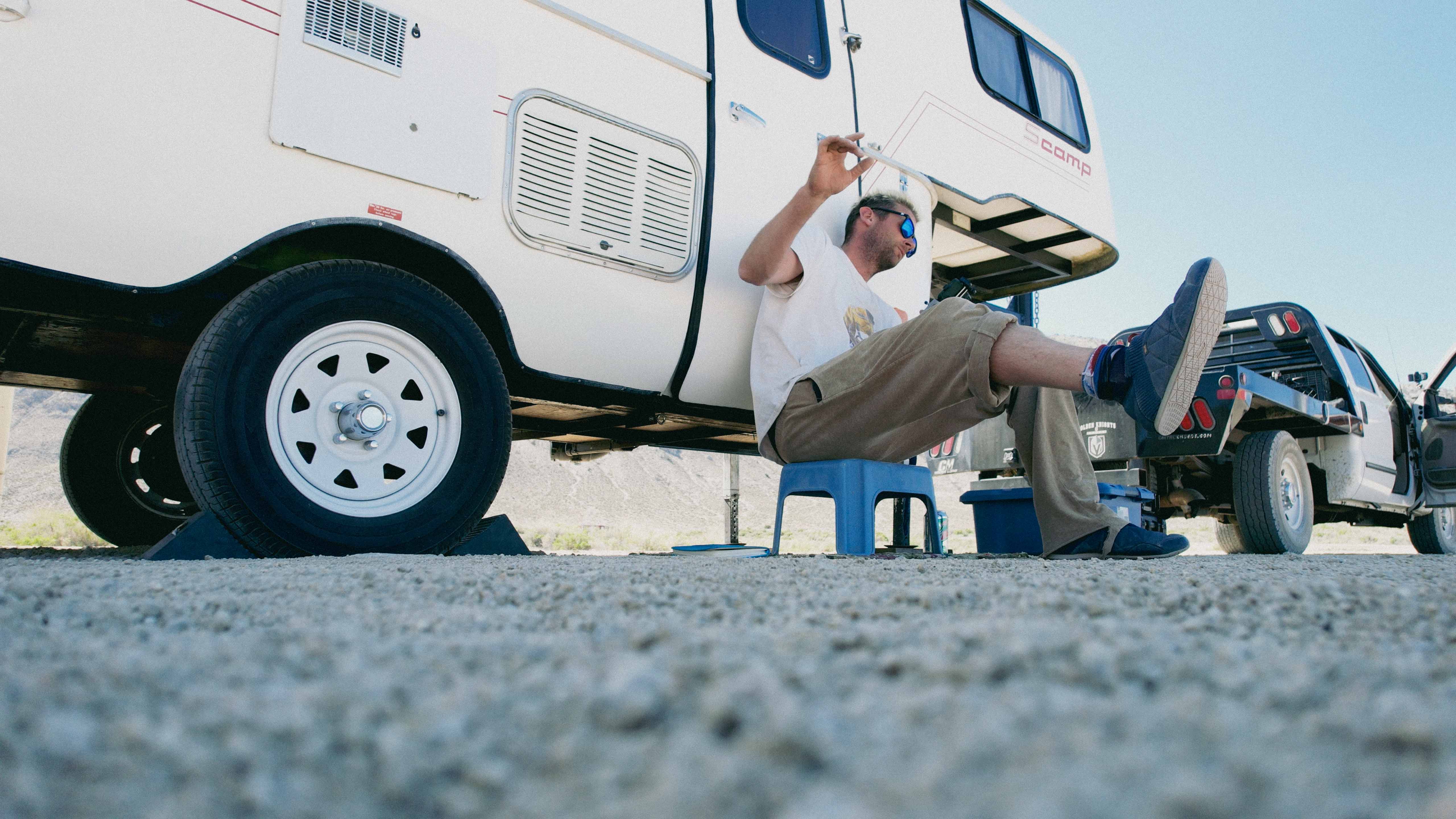 Nathan 'Mary Poppins' Altman sits near a travel trailer in the desert at a worksite, playfully dancing to music. 